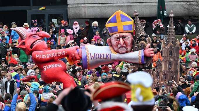Ein Mottowagen zeigt den Teufel, der am Kölner Kardinal Woelki zerrt, der sich am Kölner Dom festklammert.