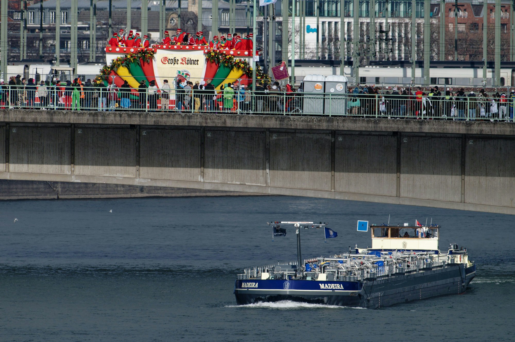 Ein Binnenschiff fährt unter der Deutzer Brücke entlang, oben ist ein Festwagen der Karnevalsgesellschaft „Große Kölner“ zu sehen.