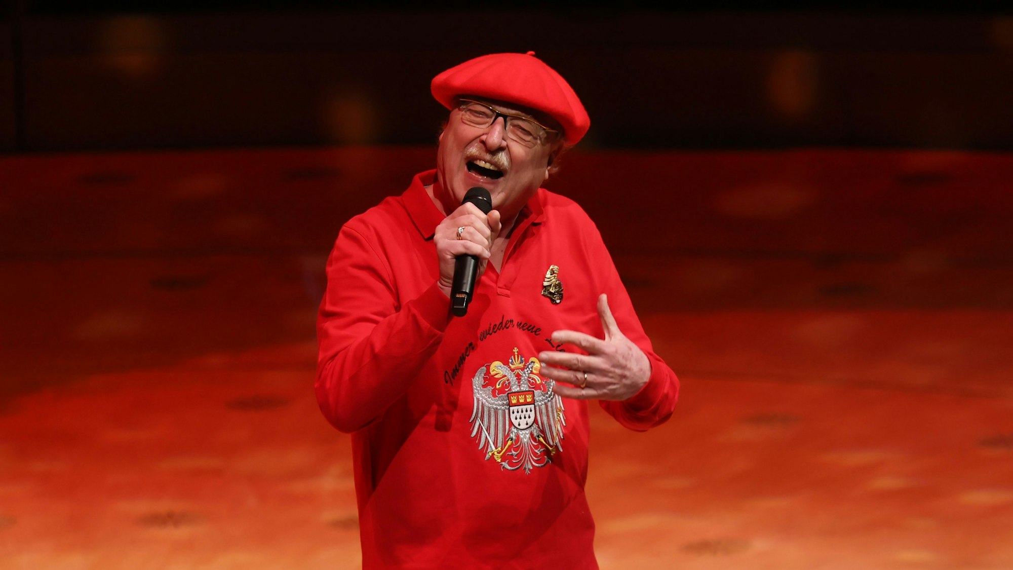 Wicky Junggeburth steht mit einem Mikro in der Hand auf der Bühne der Philharmonie Köln, er trägt einen roten Pulli mit dem Kölner Wappen drauf und eine rote Mütze.