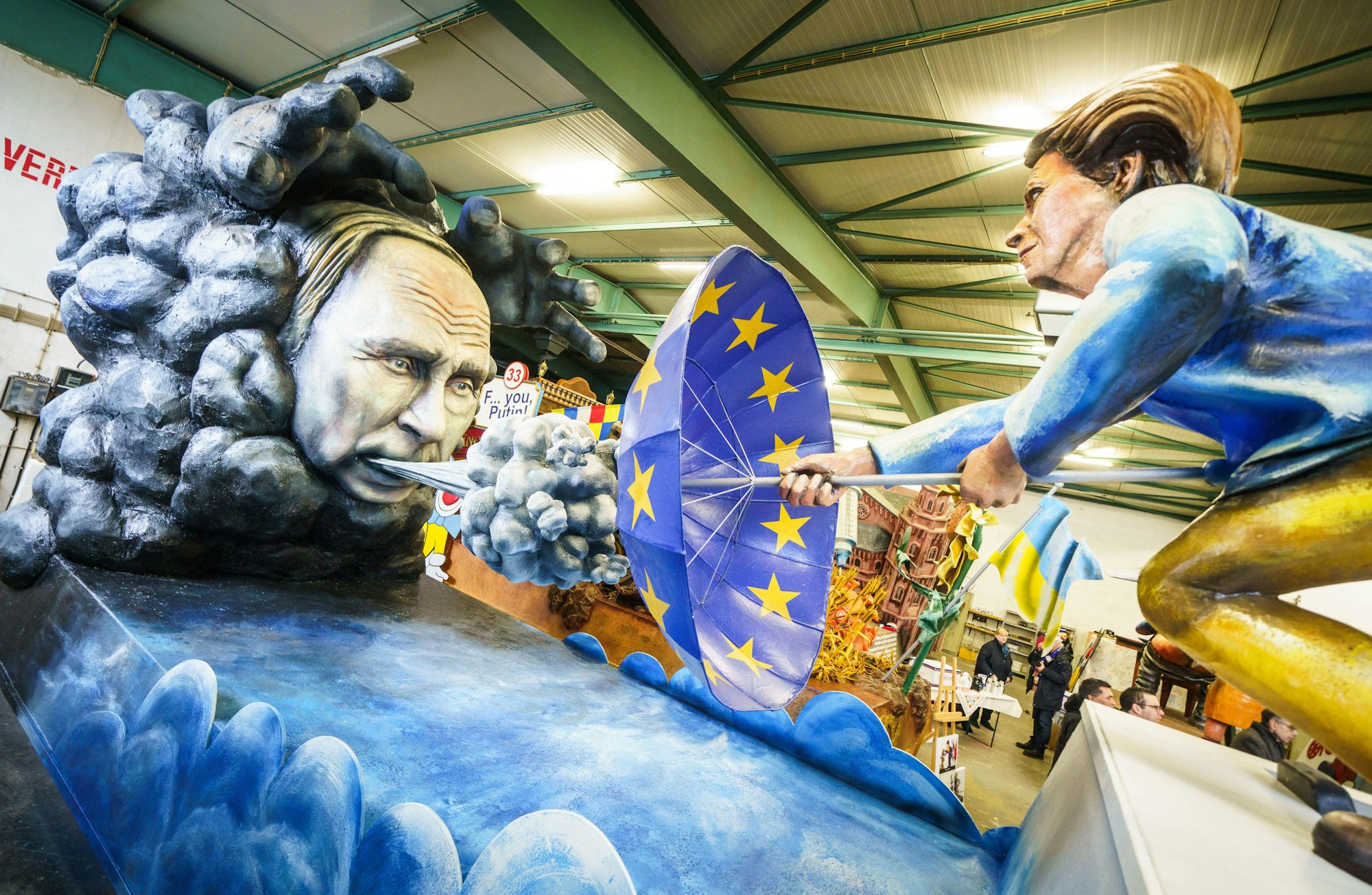«Scharfer Ostwind» heißt ein Motivwagen der Mainzer Fastnacht. Es zeigt eine Darstellung des russischen Präsidenten Putin, der Wind gegen einen Schirm in den Farben der EU-bläst, der wiederum gehalten wird von der Präsidentin der Europäischen Kommission, Ursula von der Leyen.