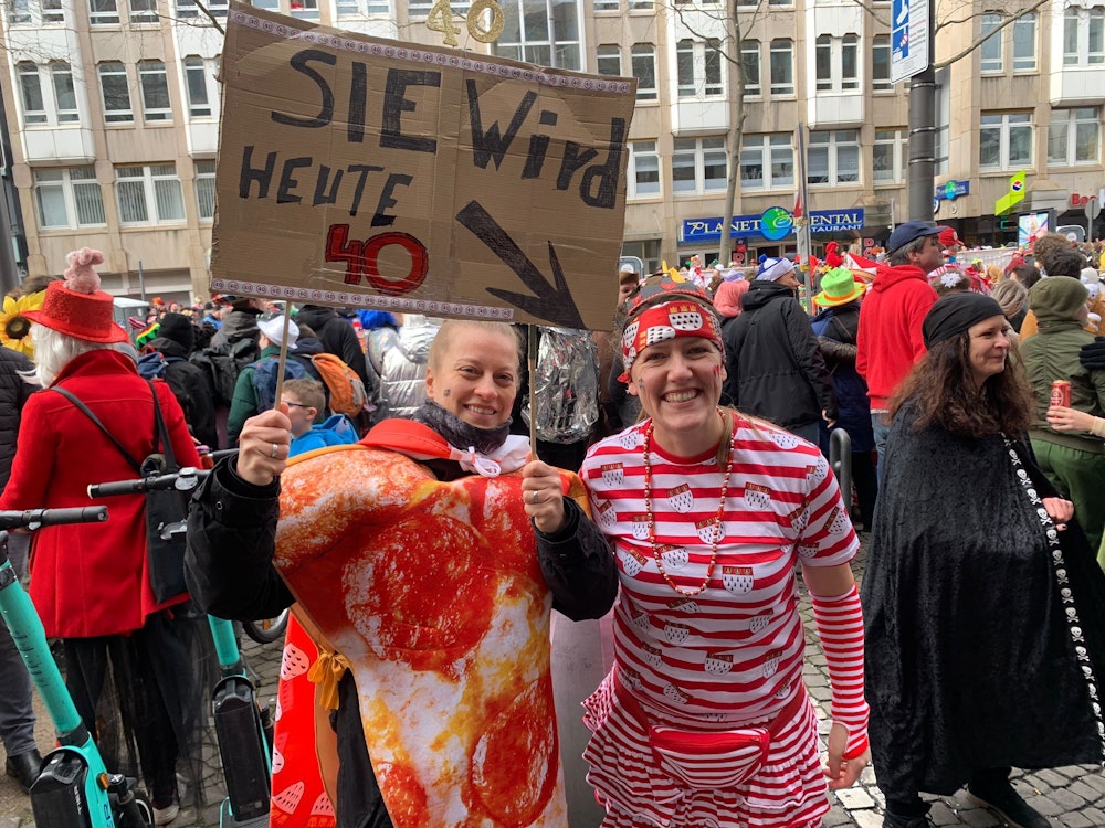 Susanne trägt ein Shirt mit Köln-Wappen, neben ihr steht ihre Freundin mit einem Pappschild, darauf steht „Sie wird heute 40“.