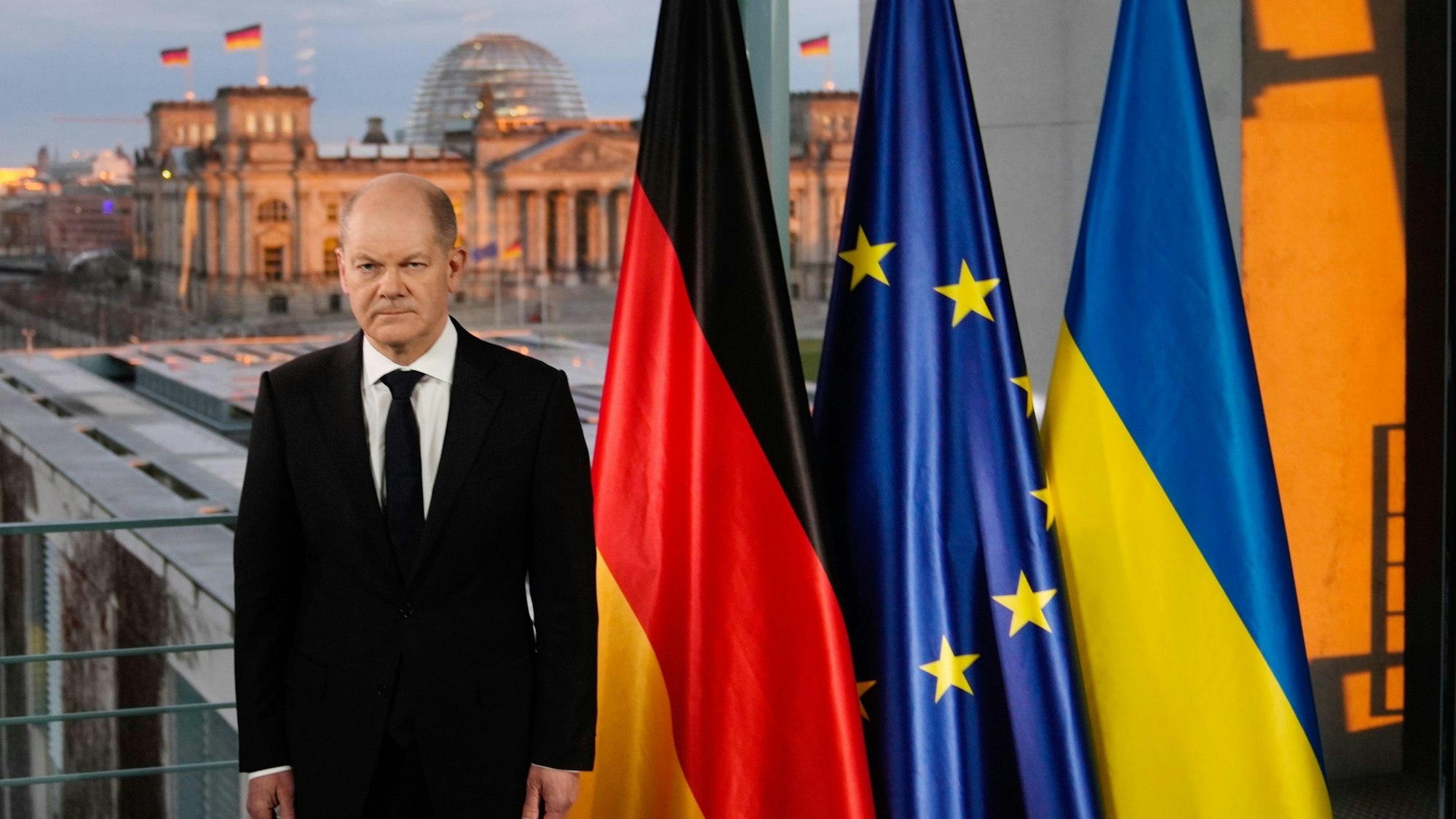Olaf Scholz steht in einem schwarzen Anzug neben einer Deutschland-, Europa- und Ukraine-Flagge.