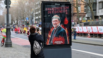 Eine Frau fotografiert das Protest-Plakat: Die Gruppe „Dies Irae“ hat an Rosenmontag diverse Werbekästen gekapert und Plakate gegen das Kölner Erzbistum und Kardinal Woelki aufgehängt.