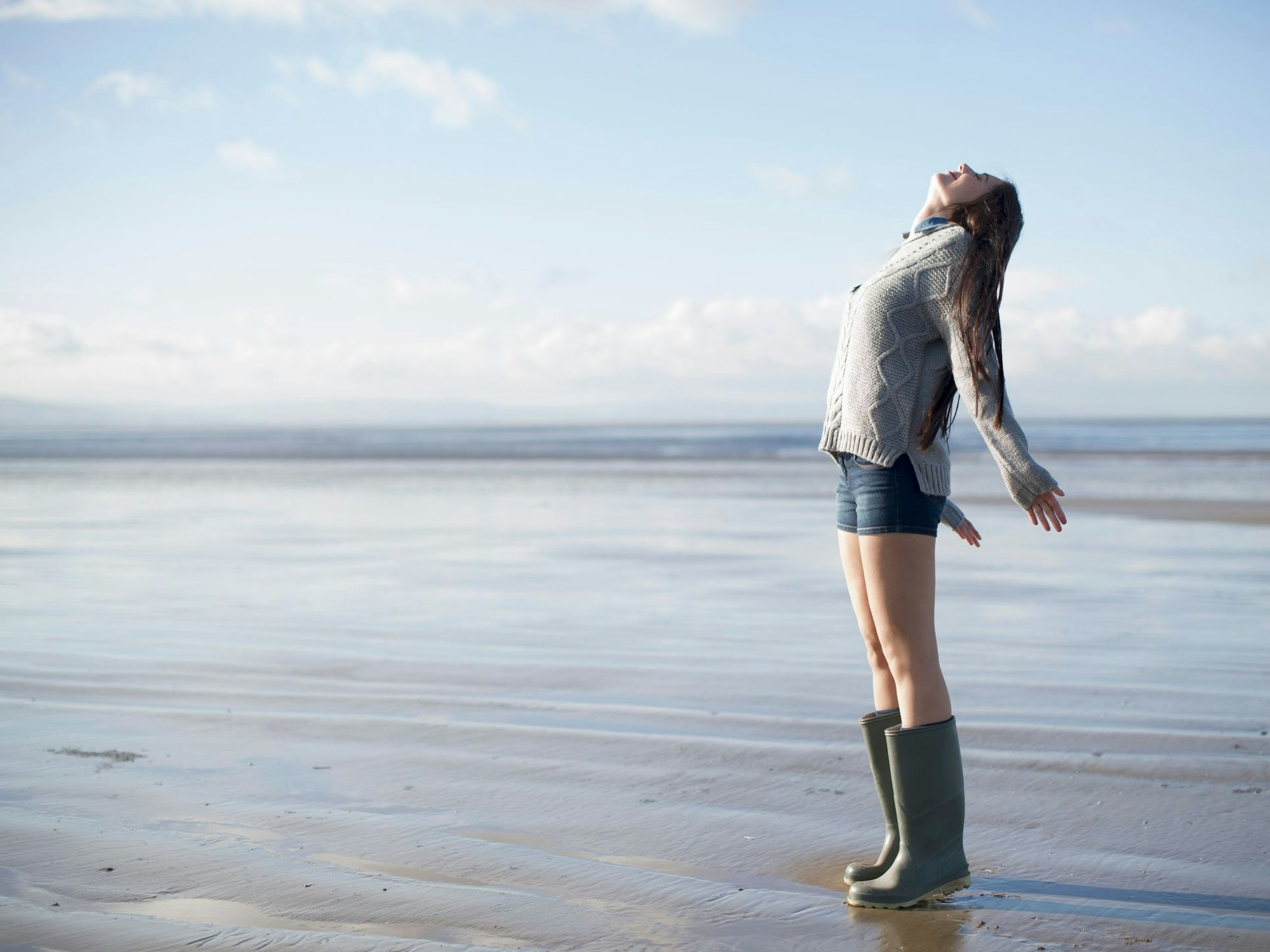 eine junge Frau in Gummistiefeln atmet am Strand tief ein
