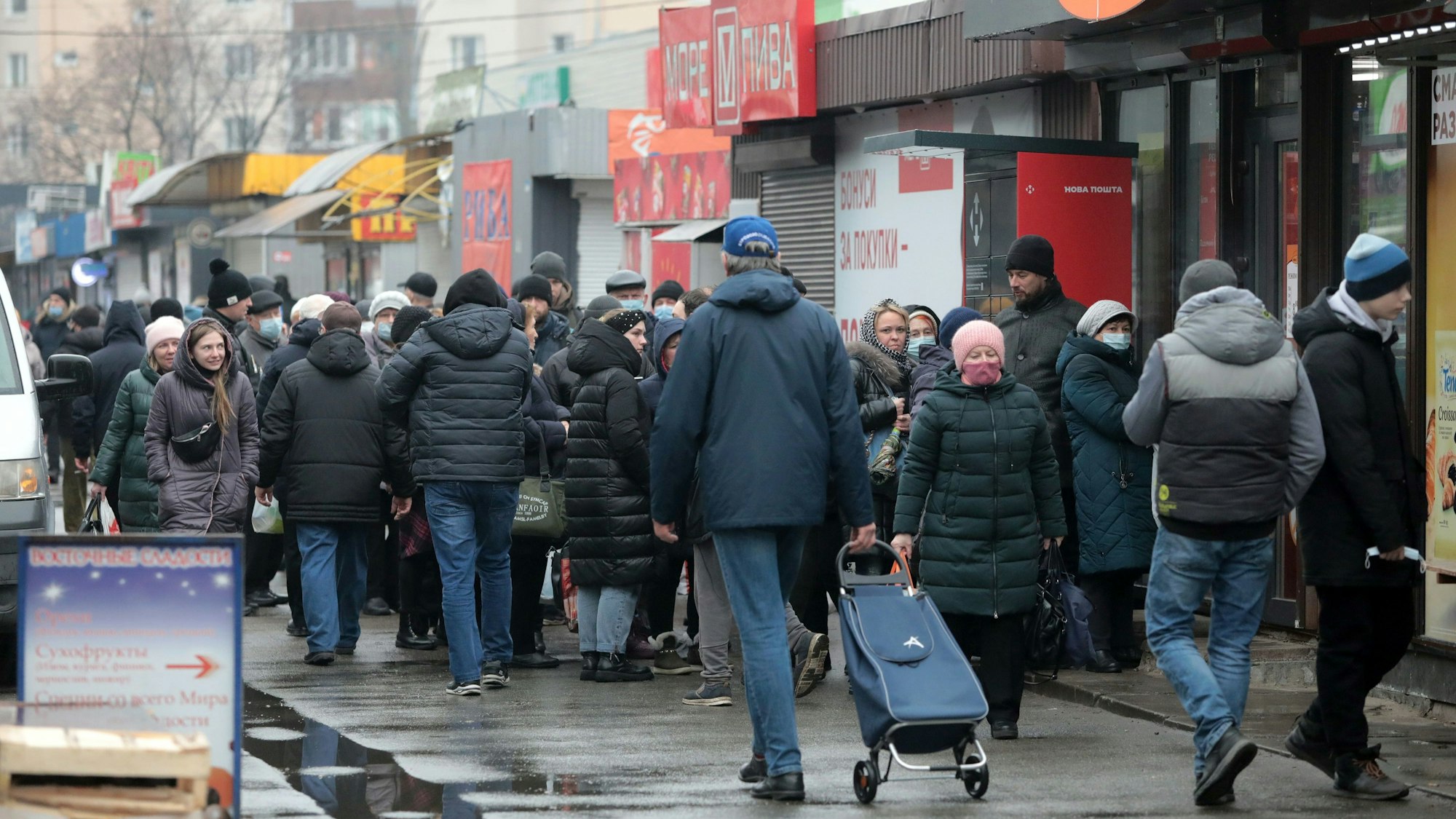 Menschen stehen in einer Schlange vor einem Lebensmittelladen. Viele haben Einkaufstüten und Taschen dabei.