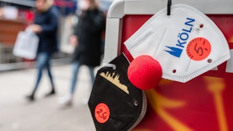 Eine FFP2-Maske mit einer roten Clownsnase hängt an einem Aufsteller vor einem Geschäft.