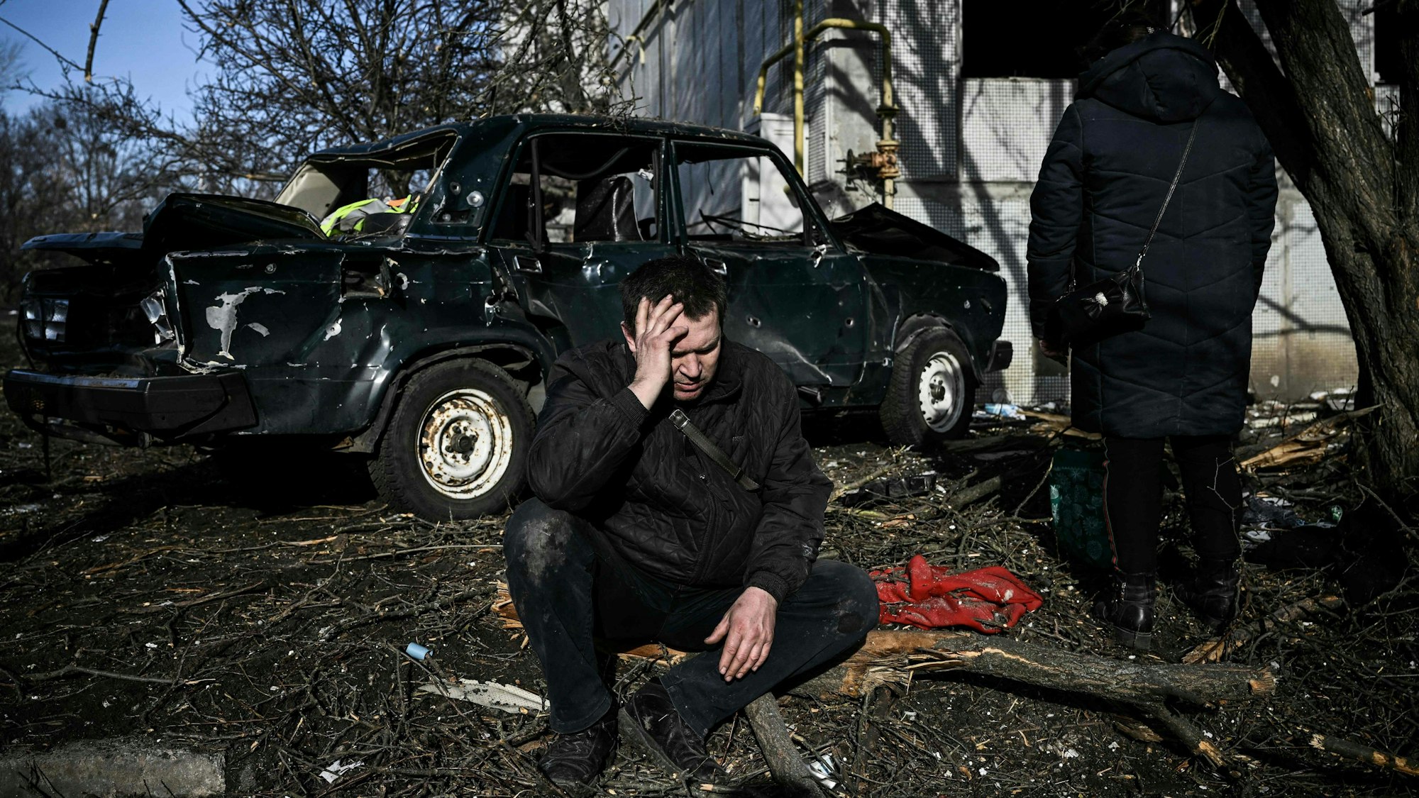 Ein Mann kniet vor einem zerstörten Auto und hält sich den Kopf.