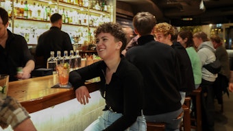 Eine junge Frau sitzt an der Bar. Vor ihr steht ein Drink, hinter ihr sitzen weitere Besucher.