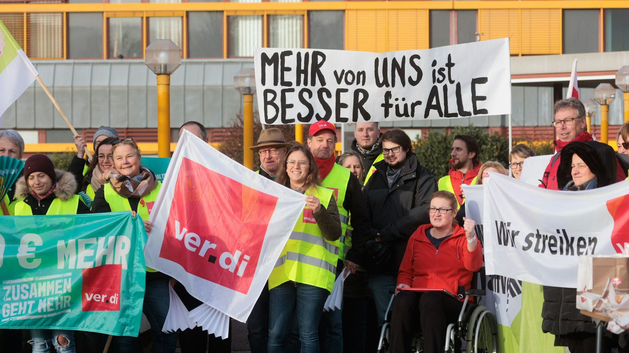 Bei einem Streik halten mehrere Personen, bekleidet mit gelben Warnwesten, Schilder und Banner in die Höhe. Darauf steht: „Wir streiken“, „Verdi“ und „Mehr von uns ist besser für alle“.