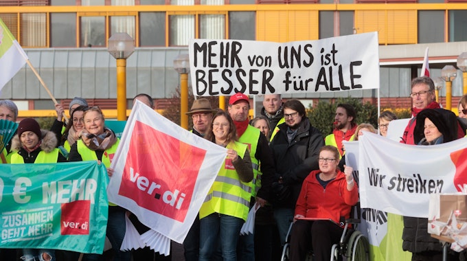 Bei einem Streik halten mehrere Personen, bekleidet mit gelben Warnwesten, Schilder und Banner in die Höhe. Darauf steht: „Wir streiken“, „Verdi“ und „Mehr von uns ist besser für alle“.