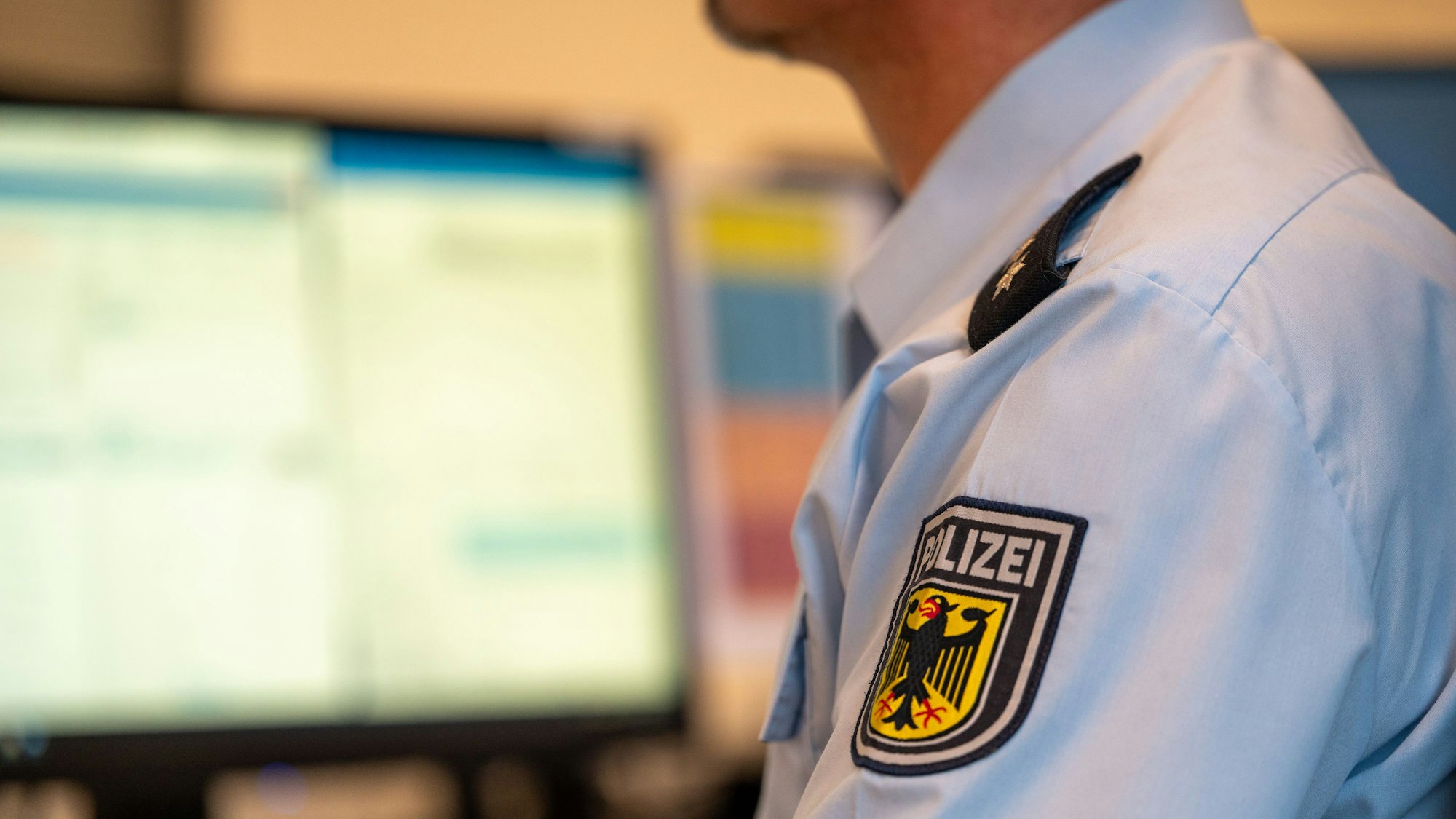 Das Symbolfoto von 2023 zeigt einen Polizeibeamten von der Seite. Er trägt ein blaues Hemd samt Polizei-Abzeichen, im Hintergrund stehen Computerbildschirme.