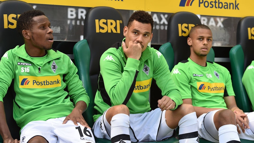 Timothee Kolodziejczak (M.) sitzt am 6. Mai 2017 auf der Ersatzbank von Borussia Mönchengladbach im Borussia-Park zwischen seinen damaligen Teamkollegen Ibrahima Traoré (l.) und Djibril Sow. Alle tragen ein grünes Trainings-Oberteil sowie weiße Hosen und Stutzen.