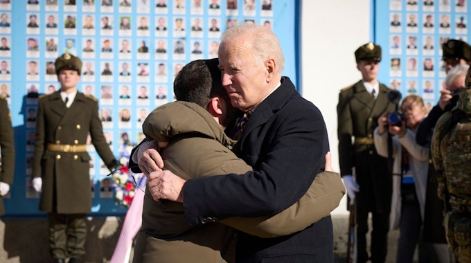 Joe Biden (Mitte r), Präsident der USA, und Wolodymyr Selenskyj, Präsident der Ukraine, umarmen sich zum Abschied an der Gedenkmauer für die gefallenen Verteidiger der Ukraine in Kiew am 20. Februar 2023. Im Hintergrund sind eine Ehrenwache ukrainischer Soldaten und ein Blumenkranz zu sehen.
