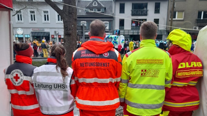 Einsatzkräfte von DRK, Johanniter, ASB und DLRG stehen nebeneinander und beobachten den Karnevalszug in Bergisch Gladbachs Stadtmitte.