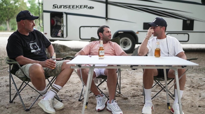 Frank Rosin, Ali Güngörmüş und Alexander Kumptner sitzen an einem Campingtisch vor einem Wohnmobil.