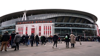 Das Emirates Stadium ist seit 2006 die Heimstätte des Premier-League-Vereins FC Arsenal. Das Foto zeigt das Stadion von außen am 11. Februar 2023, dem Tag des Arsenal-Heimspiels gegen Brentford. Fans überqueren den Vorplatz des Stadiums in Winterkleidung. 