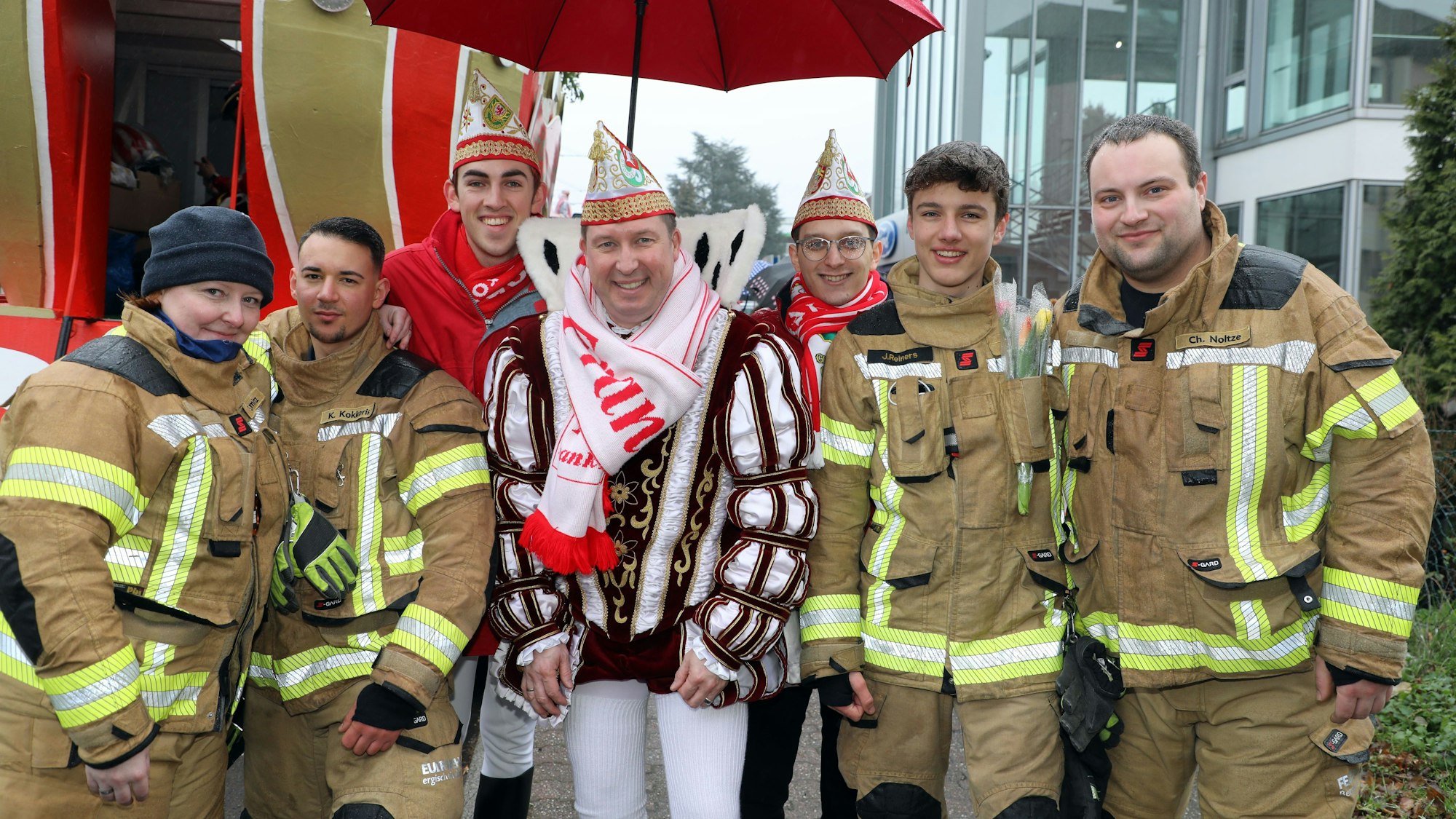 Feuerwehrleute vom Löschzug Bensberg stehen neben dem Karnevalsprinzen Frank III. (Haag), der von einem Schirm gegen den Nieselregen geschützt wird.