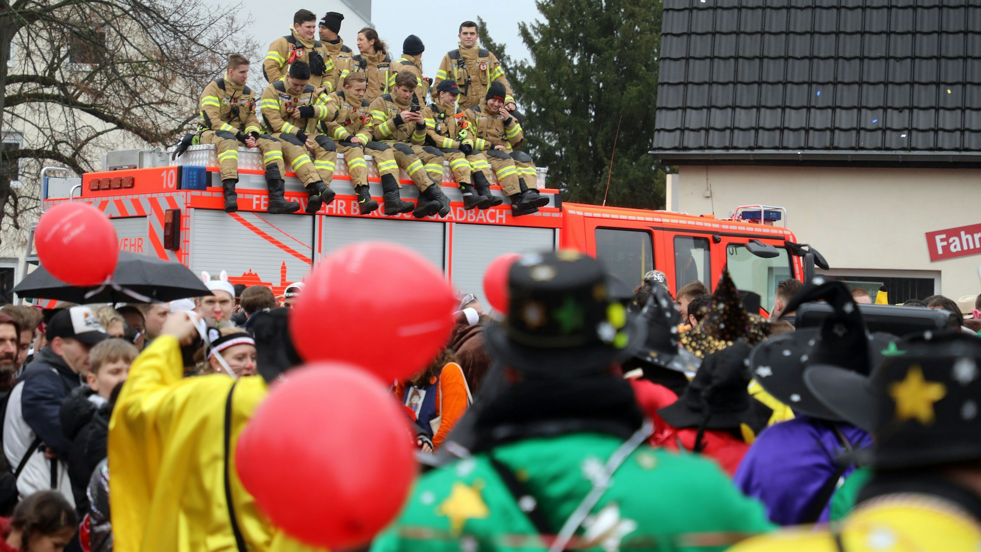 Feuerwehrleute sitzen auf dem Dach eines Feuerwehrfahrzeugs und beobachten das Karnevalsgeschehen auf der Straße.
