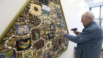 Ein Mann in blauer Jacke befestigt mit einer Klebepistole abgefallene Kieselsteine an dem großen Mosaik, auf dem Schüler die Schöpfungsgeschichte darstellten.