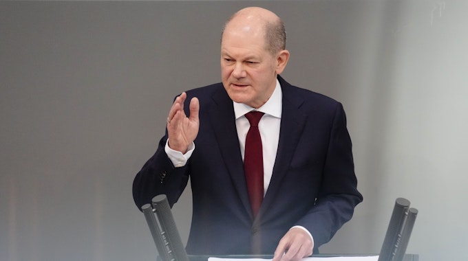 Bundeskanzler Olaf Scholz steht am Rednerpult des Bundestags. Er hat eine Hand erhoben.