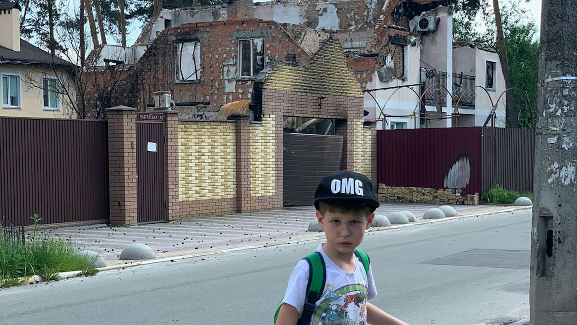 Olgas Sohn Alexander steht mit seinem City-Roller auf den Straßen von Irpin. Hinter ihm sieht man die Ruinen von zerstörten Häusern.