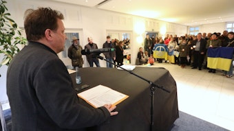 Bürgermeister Lutz Wagner steht an einem Mikrofon, das Redemanuskript liegt vor ihm. Im Saal stehen Menschen, die die ukrainische Flagge und Kerzen halten.