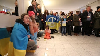 Eine Frau hat die blau-gelbe ukrainische Flagge umgehangen. Auf dem Boden stehen Windlichter. Im Hintergrund halten Menschen eine ukrainische Flagge mit dem Peace-Zeichen.