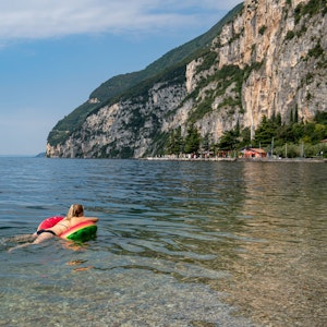 Das Symbolfoto aus dem Jahr 2021 zeigt eine Frau im Bikini, die auf einer Luftmatratze im Gardasee schwimmt. Im Hintergrund steht ein Felsmassiv.