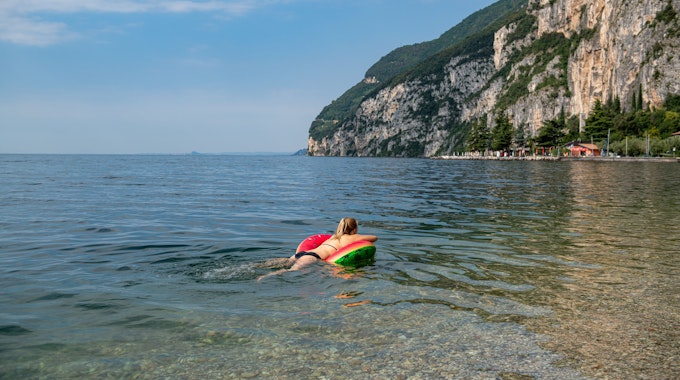 Das Symbolfoto aus dem Jahr 2021 zeigt eine Frau im Bikini, die auf einer Luftmatratze im Gardasee schwimmt. Im Hintergrund steht ein Felsmassiv.