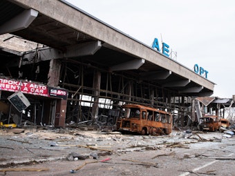 Das Foto zeigt den zerstörten Flughafen der ukrainischen Stadt Cherson. Überall liegen Metall- und Holzteile herum, das Gebäudeinnere ist bis auf das Gestellt heruntergebrannt. Einige orangefarbene Busse ohne Scheiben stehen vor dem Flughafen.