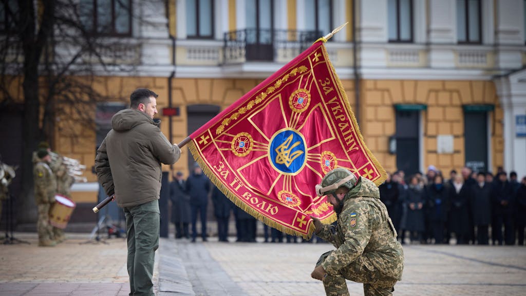 Das Foto vom 24. Februar 2023 zeigt den ukrainischen Präsidenten Wolodymyr Selenskyj. Er hält die rote Fahne einer Militäreinheit, vor ihm kniet eine Offizier in Uniform, der mit seiner rechten Hand ebenfalls die Fahne berührt.