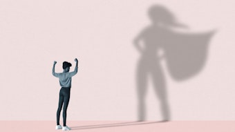 Eine junge Frau streckt ihre Arme in die Höhe, vor einer rosafarbenen Wand, auf der der Schatten einer Superwoman erscheint.
