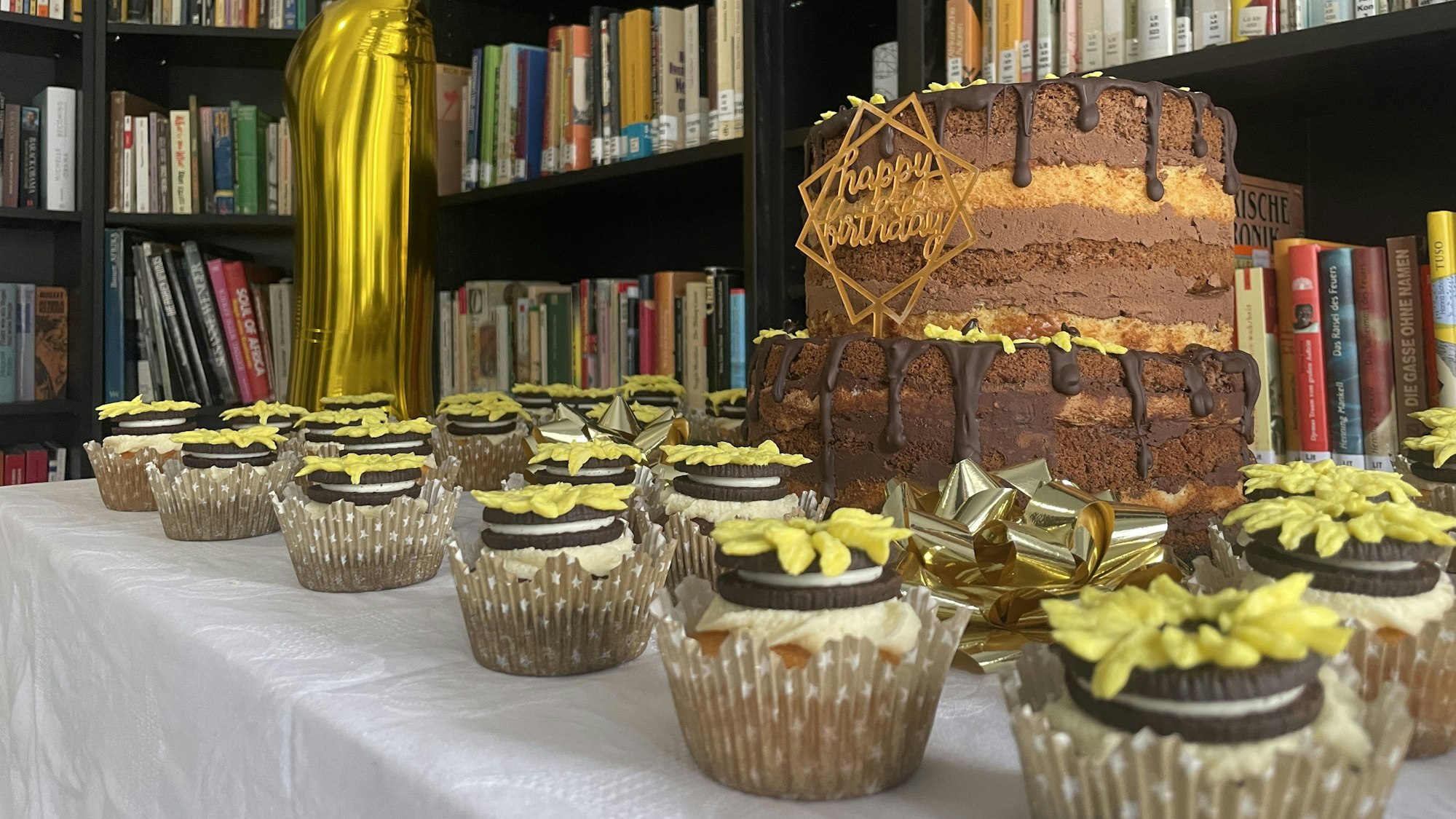 Cupcakes, die mit Sonnenblumen verziert sind, stehen um eine große Schokoladentorte. Dahinter ist ein Luftballon in der Ziffer „1“, das vor einem Bücherregal steht.