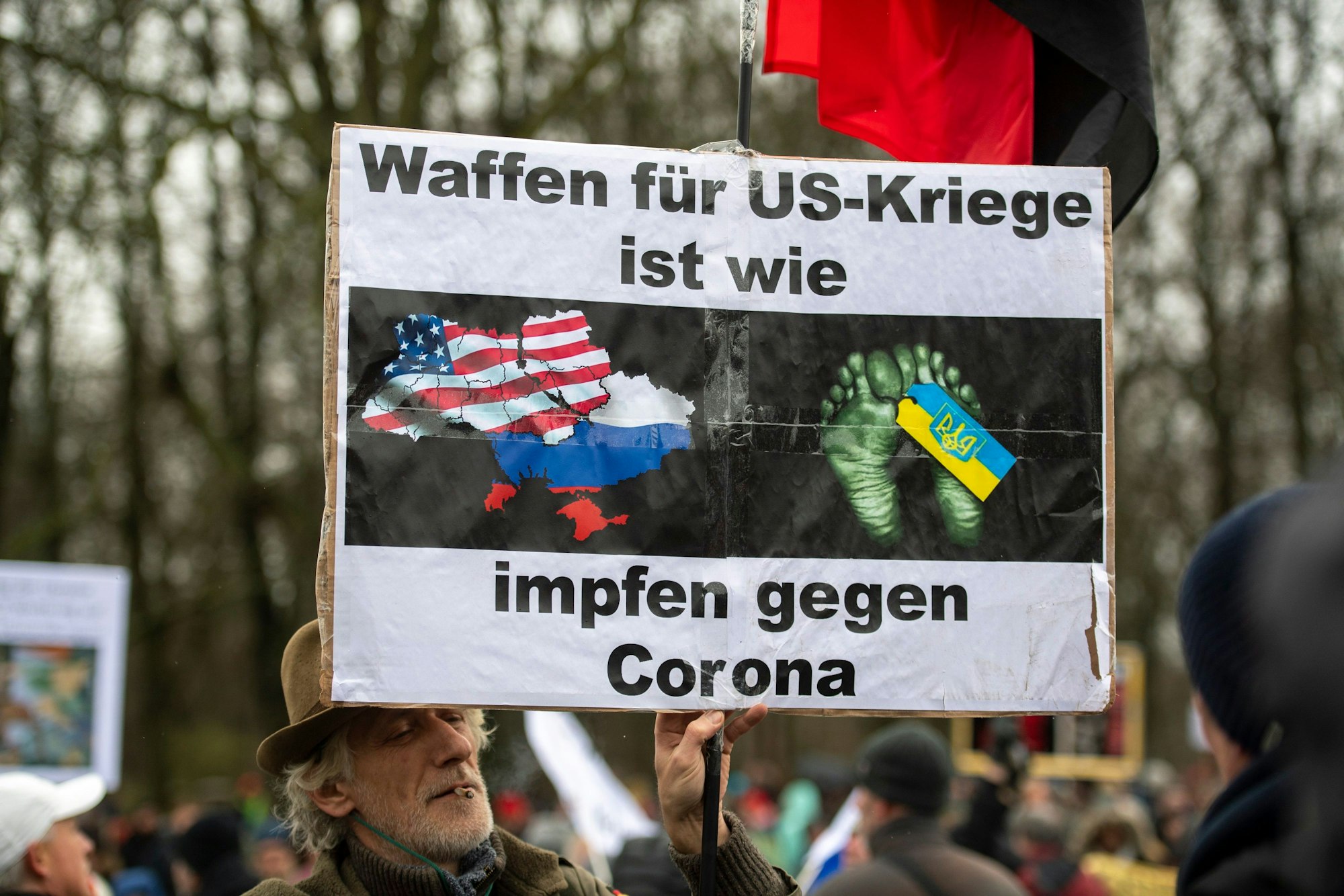 Ein Teilnehmer einer Demonstration für Verhandlungen mit Russland am Brandenburger Tor trägt ein Plakat mit der Aufschrift „Waffen für US-Kriege ist wie impfen gegen Corona“.