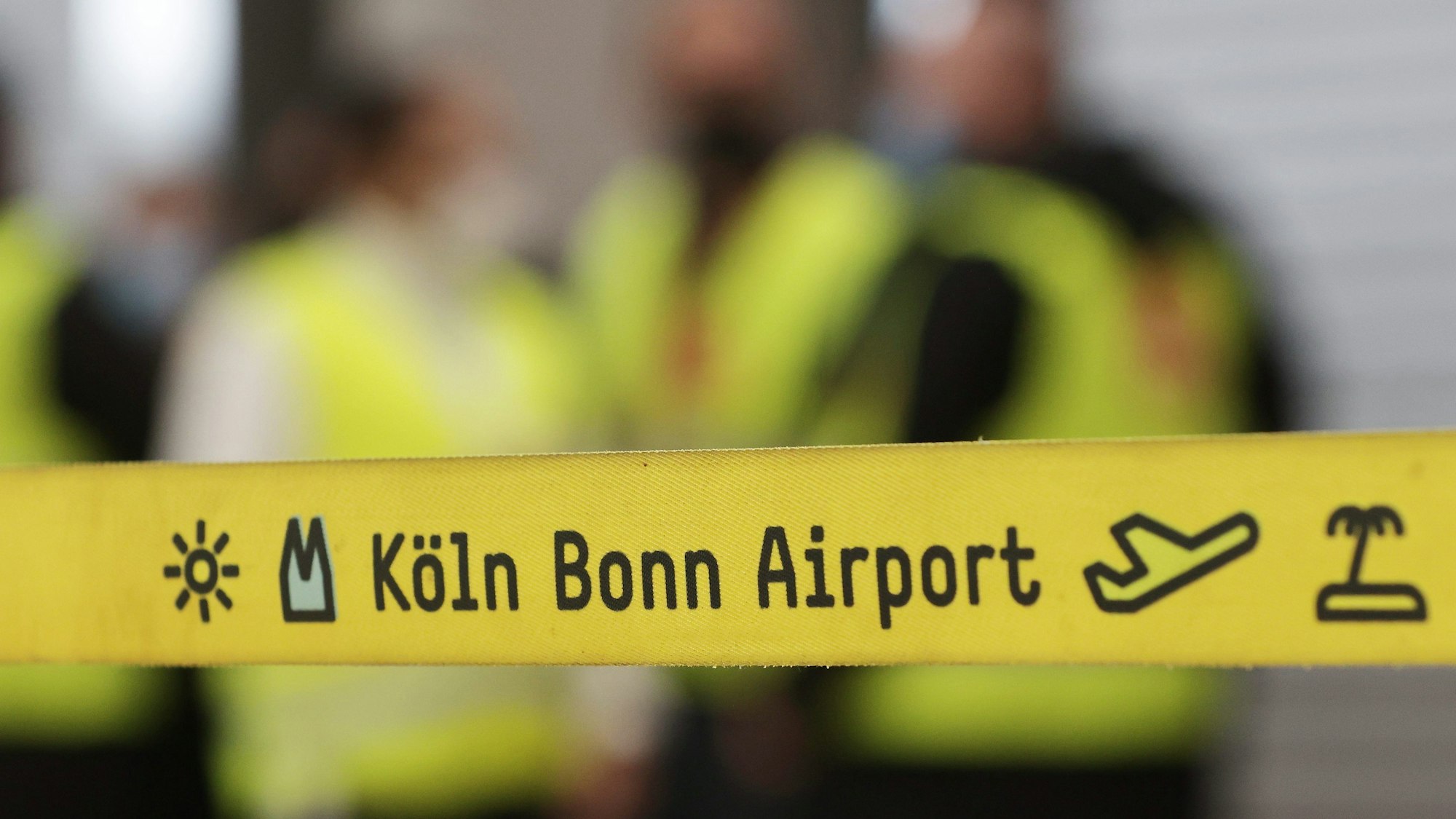Ein gelbes Absperrband mit der Aufschrift „Köln Bonn Airport“ hängt vor eine Gruppe Streikender während eines Warnstreiks.