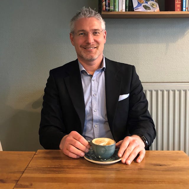 Martin Sagel aus Kerpen sitzt im Anzug in einem Café. In seinen Händen hält er einen Cappuccino.
