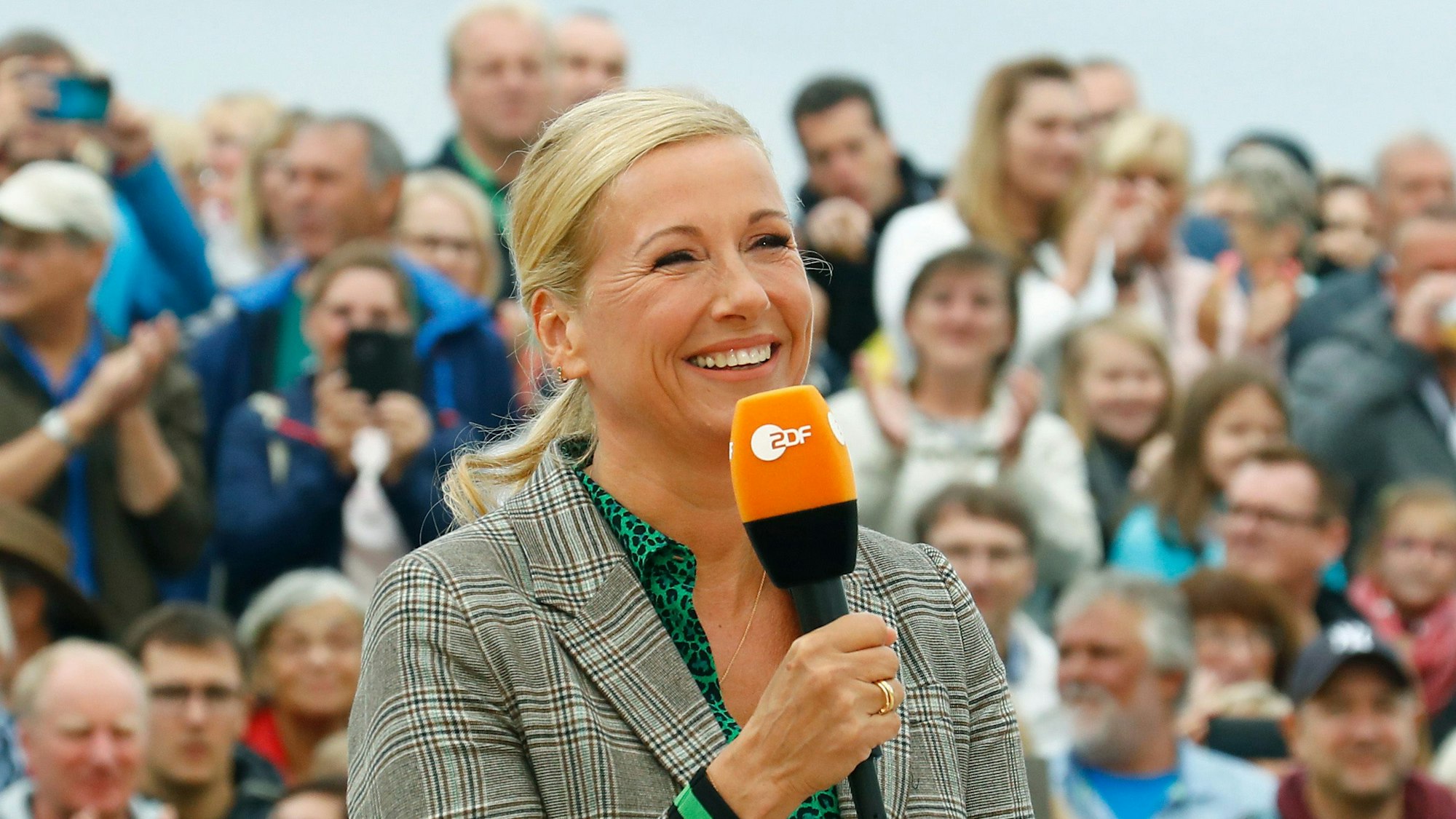 Andrea Kiewel (57) bei der Moderation des „ZDF-Fernsehgartens“. Die Moderatorin hält ein Mikrofon mit dem Senderlogo in der Hand, im Hintergrund ist das Publikum zu sehen.