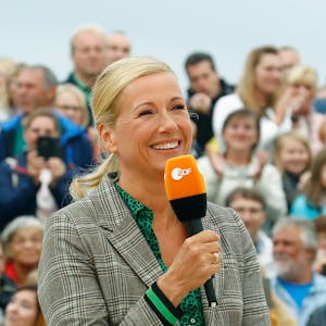 Andrea Kiewel (57) bei der Moderation des „ZDF-Fernsehgartens“. Die Moderatorin hält ein Mikrofon mit dem Senderlogo in der Hand, im Hintergrund ist das Publikum zu sehen.&nbsp;