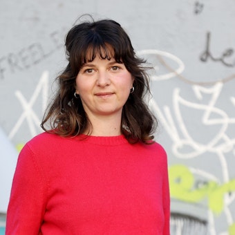 Leonie Pfennig trägt einen pinken Pullover und steht vor einer Wand mit Graffitis.