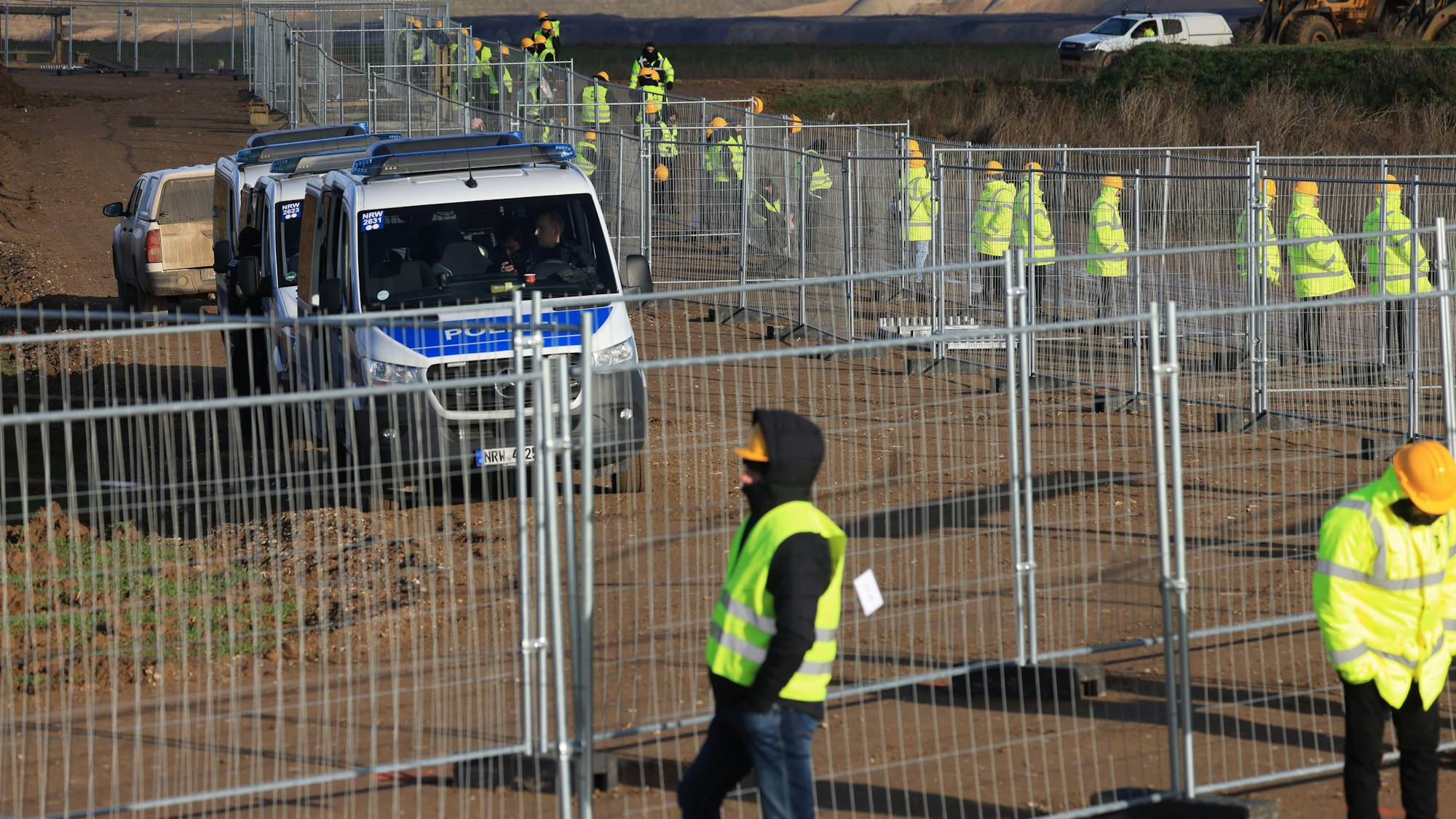 Mitarbeiter einer Werkschutzfirma stehen im Januar an einem Zaun beim von Klimaaktivisten besetzten Braunkohleort Lützerath. Sie alle tragen neongelbe Warnjacken. Hinter dem Zaun sind Vans der Polizei zu sehen.