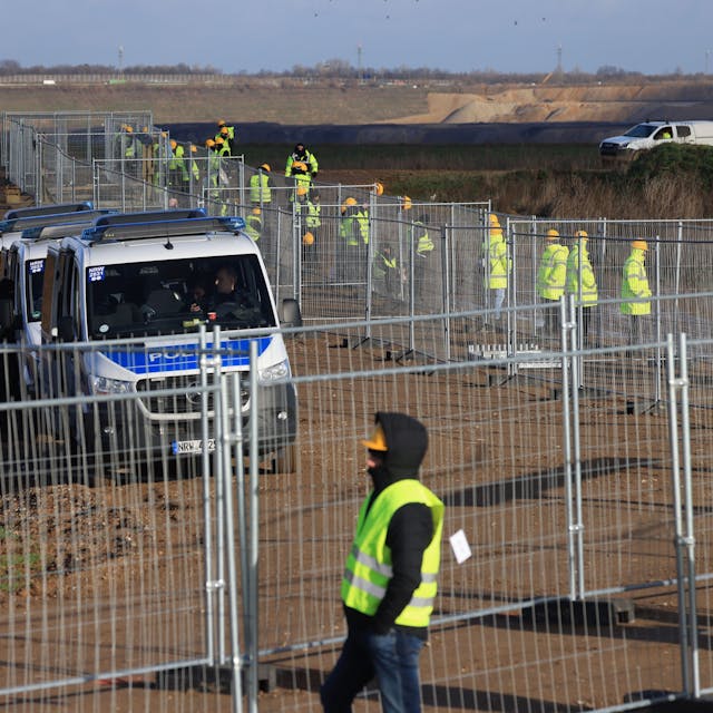Mitarbeiter einer Werkschutzfirma stehen im Januar an einem Zaun beim von Klimaaktivisten besetzten Braunkohleort Lützerath. Sie alle tragen neongelbe Warnjacken. Hinter dem Zaun sind Vans der Polizei zu sehen.&nbsp;
