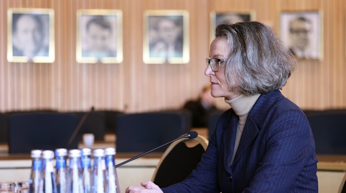 Ministerin Ina Scharrenbach (CDU) ist im Halbprofil zu sehen; sie sitzt am Mikrofon, trägt einen dunkelblauen Blazer und einen beigen Rollkragenpullover. Sie schaut ihr Gegenüber konzentriert an.