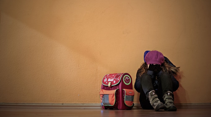 Ein Mädchen sitzt neben ihrem Schulranzen auf dem Boden und hält die Hände vor das Gesicht (Symbolfoto).
