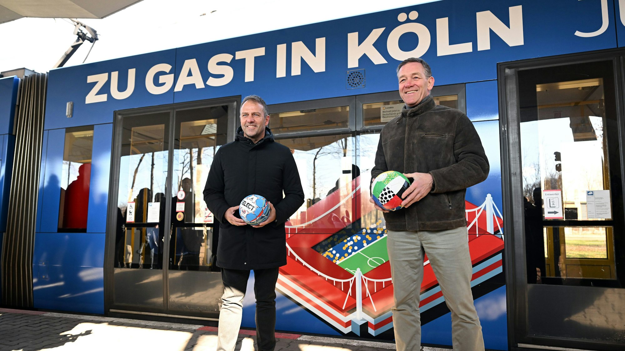 Fußball-Bundestrainer Hansi Flick und Handball-Bundestrainer Alfred Gislason stehen vor einer Straßenbahn.