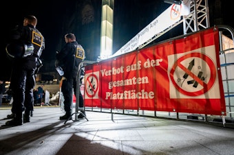 Ein Banner mit dem Hinweis auf Feuerwerksverbot an einem Absperrgitter. Daneben stehen zwei Polizisten.