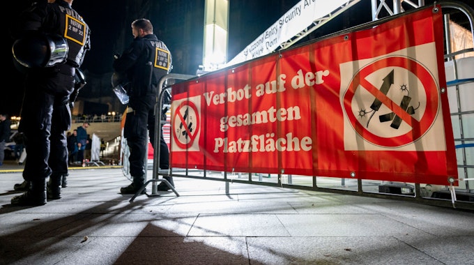 Ein Banner mit dem Hinweis auf Feuerwerksverbot an einem Absperrgitter. Daneben stehen zwei Polizisten.
