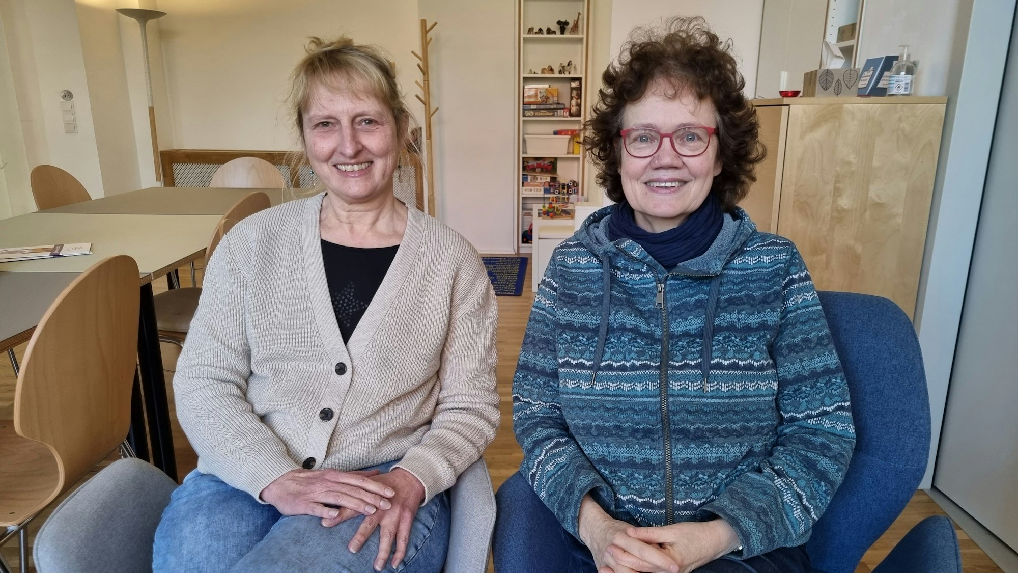 Ute Fingaß und Claudia Schrimpf sitzen auf zwei Sesseln in der Geschäftsstelle von „Frauen helfen Frauen“ in Köln und gucken in die Kamera.