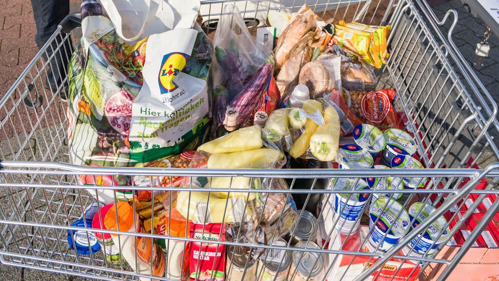 Ein Mann steht nach seinem Einkauf bei einem Discounter neben seinem vollen Einkaufswagen und dem bereits mit Lebensmitteln gut gefüllten Kofferraum seines Autos.