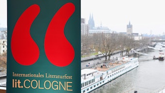 Ein Schild mit dem Logo der lit. Cologne am Rheinufer mit Blick auf den Dom.