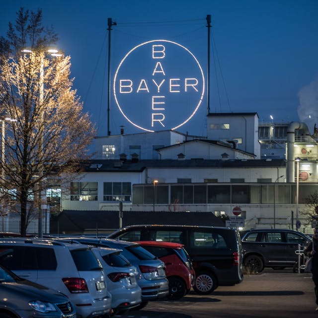 Das leuchtende Bayer-Kreuz ist aus einiger Entfernung vor parkenden Autos im Chempark Leverkusen abgebildet. Schornsteine qualmen im rechten Teil des Bildes, links befinden sich zwei Bäume.&nbsp;